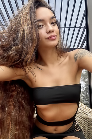 Dulce sits in silk on wicker, black lingerie against skin, teasing with limbs spread